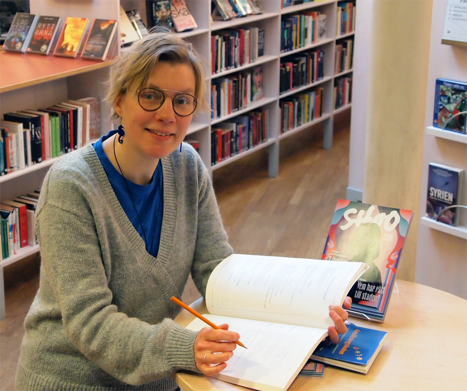 En kvinna står vid ett bord med en bok och håller i en penna.